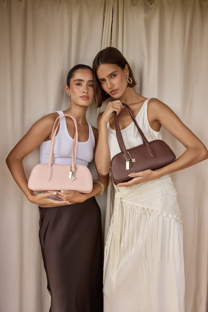 Two women pose side by side, each holding a structured handbag: blush pink on the left and brown on the right, in front of neutral curtains.