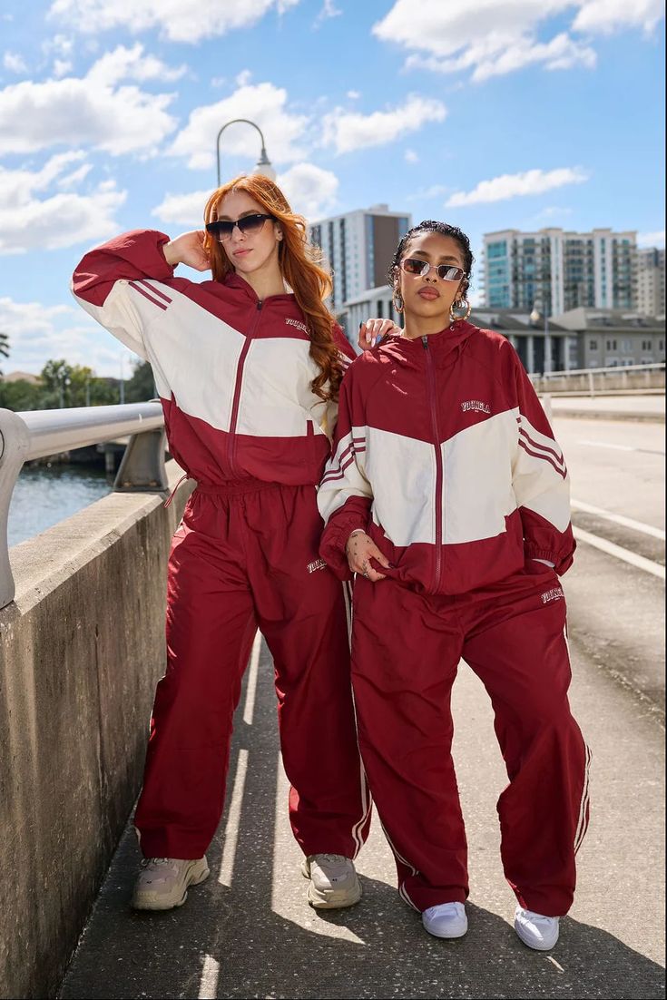 dress cool - Two women in matching maroon and white tracksuits pose on a sunny city sidewalk by a waterway, with buildings in the background.