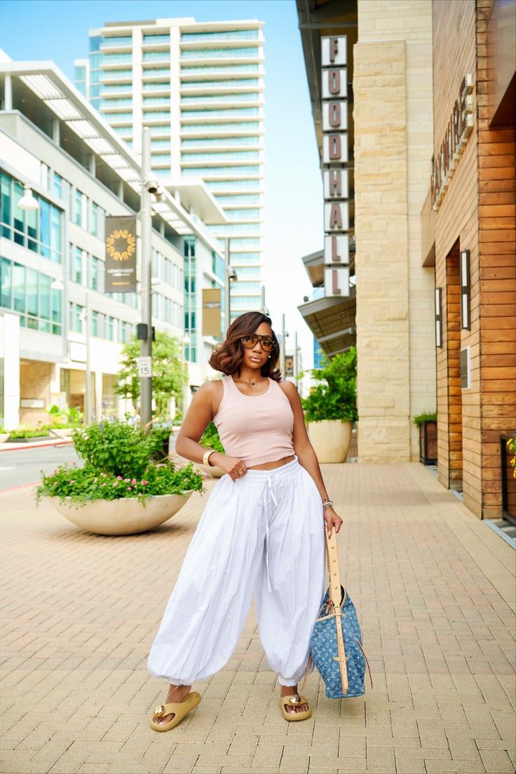 dress cool - Woman posing on an urban sidewalk in sunny day, wearing a pink crop top, white wide-leg pants, sunglasses, and carrying a blue tote bag with tan sandals.