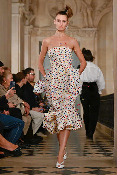 ‎A model walked the runway for Jacquemus’ Fall/Winter 2026 collection, “Le Palmier,” wearing a strapless, flared midi dress embellished with multicolored polka dots and carrying a matching purse.