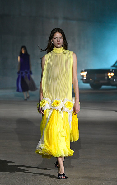 Model Kaja Krawczyk walked the runway at Dior’s 2027 Cruise collection show, wearing a high mock-neck gown featuring a pleated tunic bodice, three-dimensional fabric flowers positioned along the knees, and a floor-length tulle skirt.