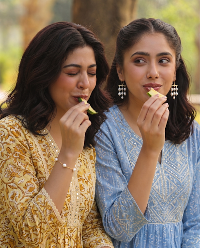 Two women in patterned dresses share a celery snack outdoors, one smiling with eyes closed while tasting a stalk.