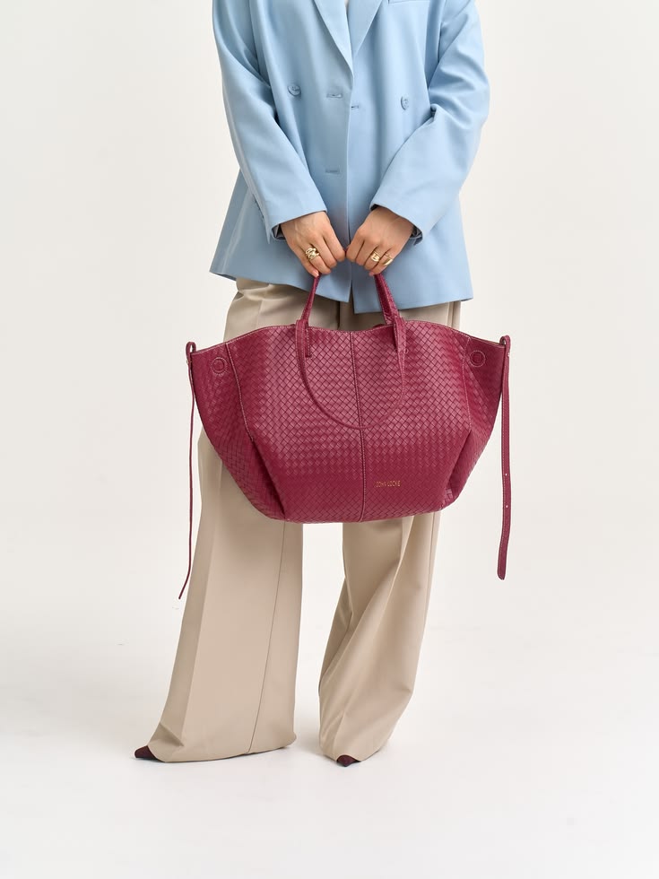 Magenta woven tote bag with handles held in front by a person wearing a light blue blazer and beige wide-leg pants.