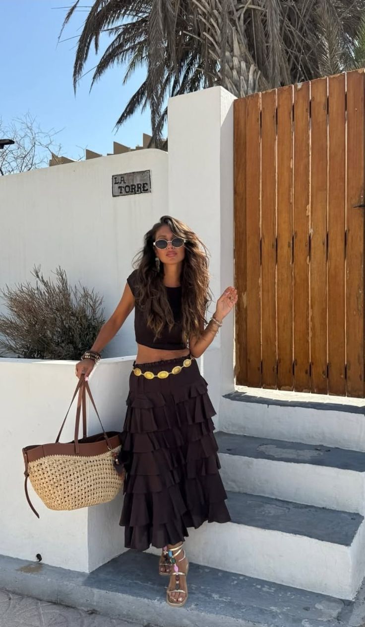 Boho chic - Woman in a black cropped top and tiered dark skirt poses on white stairs, holding a straw tote bag and wearing sunglasses with long wavy hair.