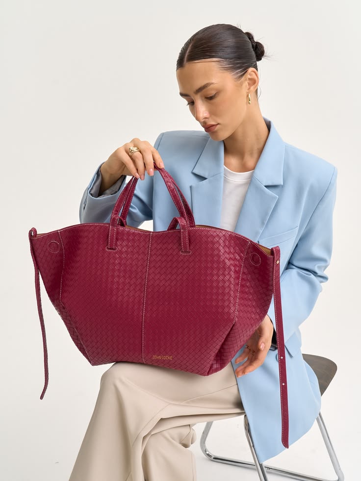 Woman in a light blue blazer sits holding a large burgundy woven tote bag, showcasing it as a fashion accessory, against a neutral backdrop.