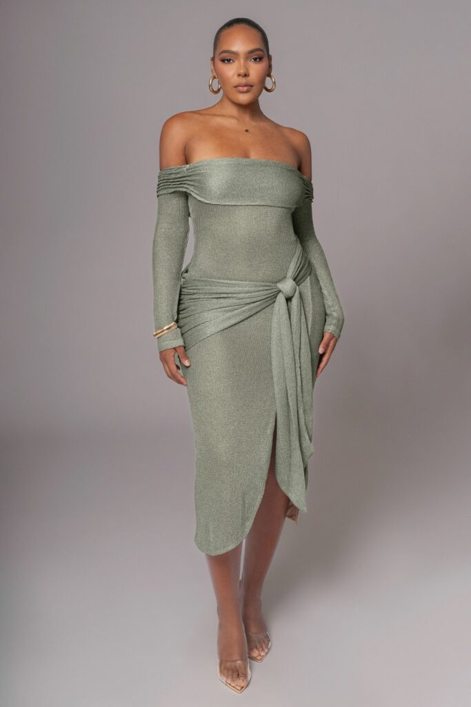Woman wearing an off-the-shoulder, metallic sage green dress with long sleeves and a front waist knot, posing against a gray backdrop.