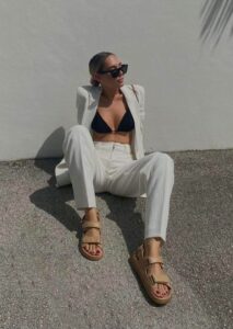 Woman in a white suit with a black bikini top sits on a gravel surface against a pale wall, wearing sunglasses and tan Chunky Sandals.