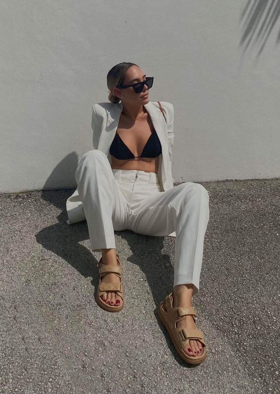 Woman in a white suit with a black bikini top sits on a gravel surface against a pale wall, wearing sunglasses and tan Chunky Sandals.