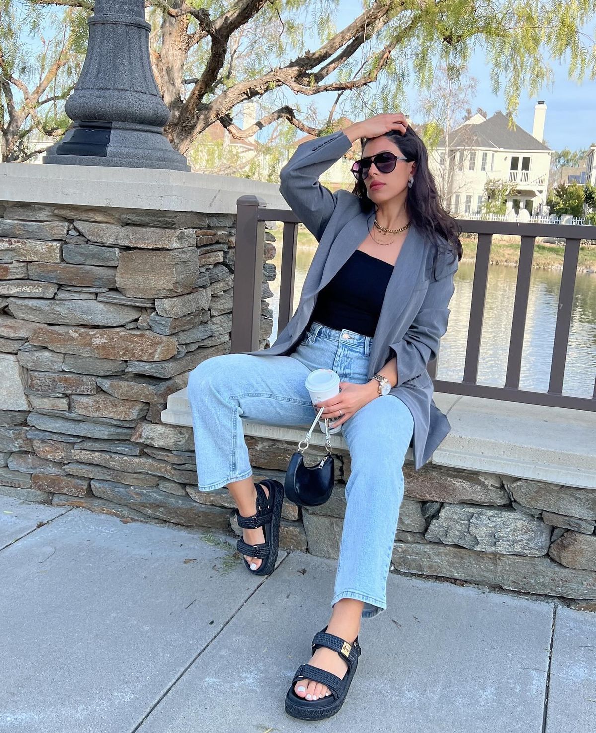 Woman sitting on a stone ledge by a railing, wearing a gray blazer, black top, light denim jeans, Chunky Sandals, sunglasses, and holding a coffee cup outdoors by the water.