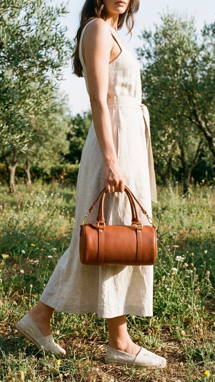 Woman in a light beige midi dress walking in a meadow, carrying a brown leather Barrel Bags and wearing matching espadrille shoes.