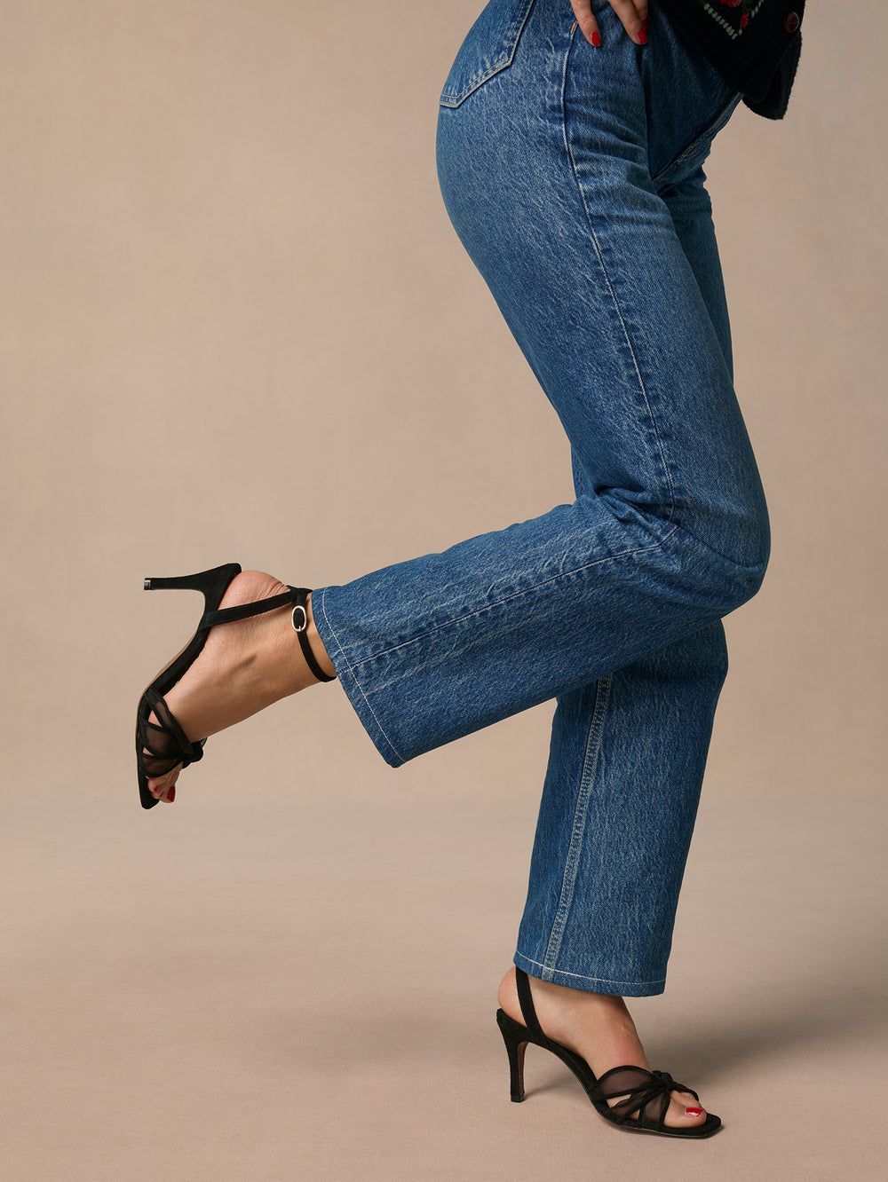Legs in blue jeans with black strappy high-heeled Bare Sandal, one foot raised as if dancing or stepping forward against a beige background.