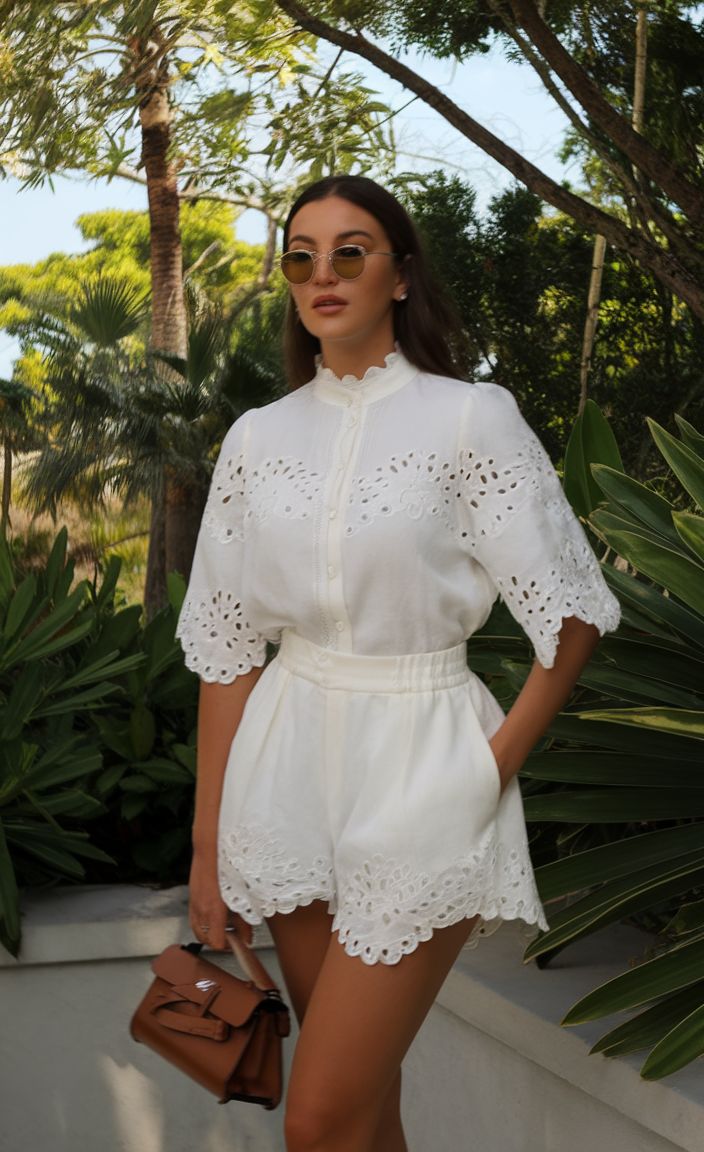Woman in White eyelet rompers standing outdoors among tropical plants, wearing round sunglasses and carrying a brown handbag.