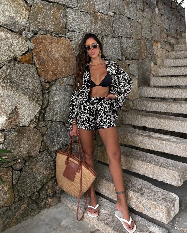 Woman in a Matching Neutral Set Trends black bikini top with a black-and-white patterned set, sunglasses, and a woven straw bag posing by a stone wall and steps.