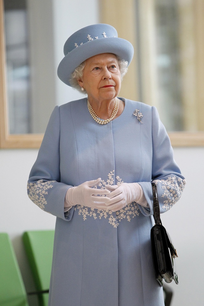 Elderly woman in a pale blue coat and matching hat with pearl necklace and gloves, holding a black handbag indoors.