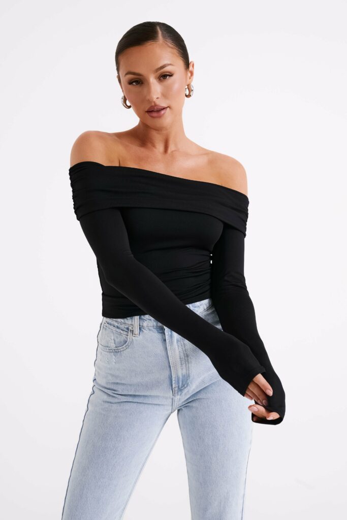 Woman wearing a black off-the-shoulder top and light-wash jeans, posed against a white background with arms crossed in front.