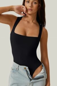 Model wearing a black square-neck bodysuit with light-wash jeans, posing with one hand near her face in a studio.