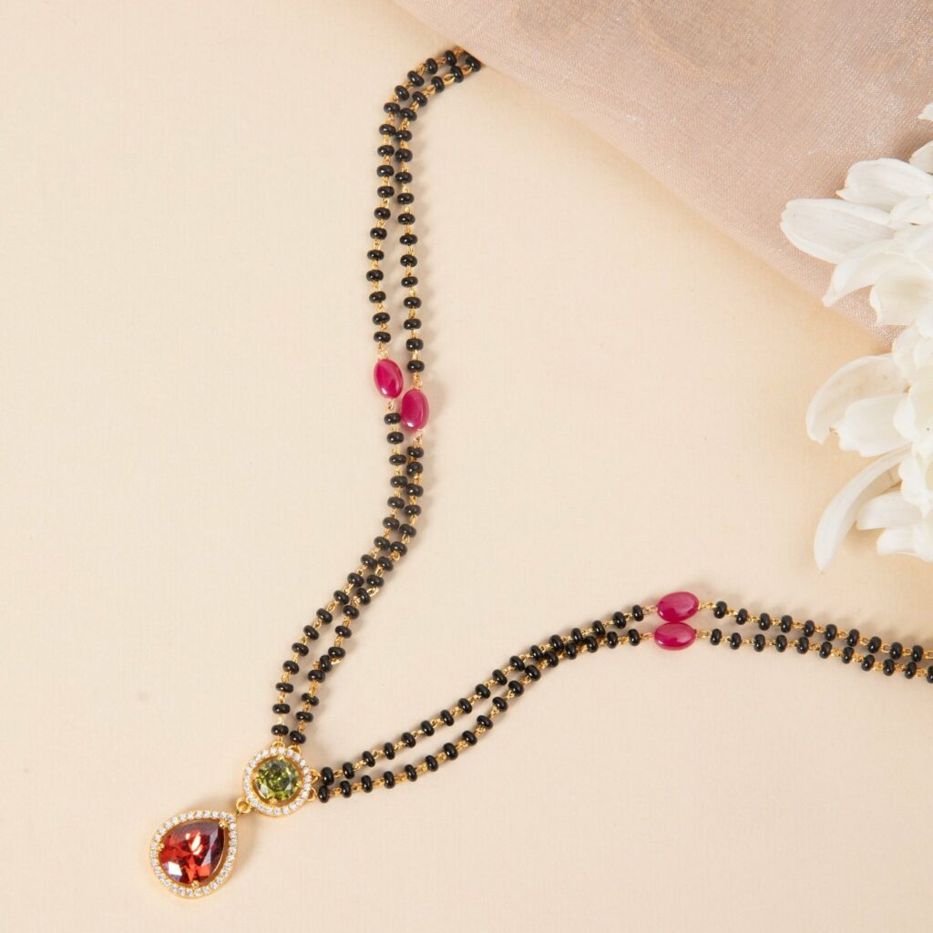 Gold and black beaded necklace with red teardrop and green centerpiece pendants on a beige surface.