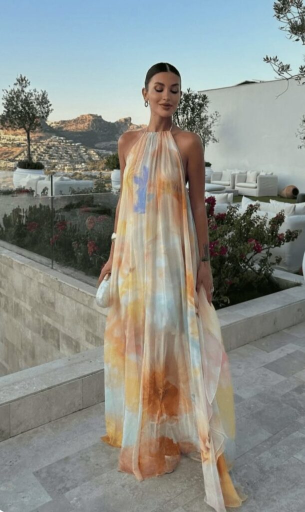 Woman with slicked hair wearing a pastel tie-dye halter gown, posing on a terrace with a scenic hillside view in the background, holding a small white clutch.