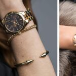 Two wrists display stylish jewelry: a gold-tone wristwatch with matching bracelets on dark fabric, left side close-up.