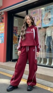 Young woman in a burgundy tracksuit with white stripes, standing on a city sidewalk in front of a storefront window.