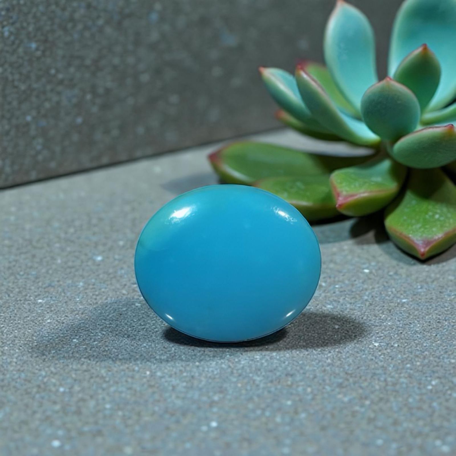 December Birthstone Blue glossy sphere resting on a gray speckled surface with a blurred succulent plant in the background.