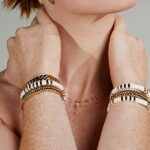 Close-up of a person’s neck and wrists adorned with gold and white bracelets and a delicate necklace with a cross, against a neutral backdrop