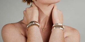 Close-up of a person’s neck and wrists adorned with gold and white bracelets and a delicate necklace with a cross, against a neutral backdrop