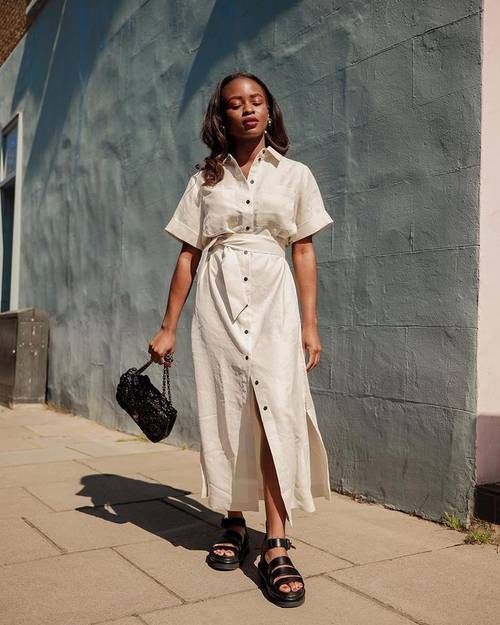 Woman in a white shirt dress with a belt walks along a sunny sidewalk, carrying a small black purse and wearing black Chunky Sandals.