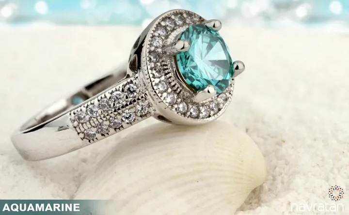 Silver ring with a round aquamarine pendant stone surrounded by diamonds in a halo setting, resting on a sandy surface.