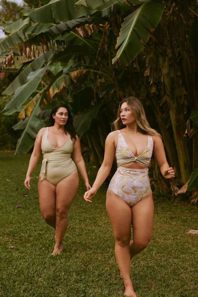 Two women in One-Piece Swimsuits walk barefoot on grass beneath large tropical leaves.