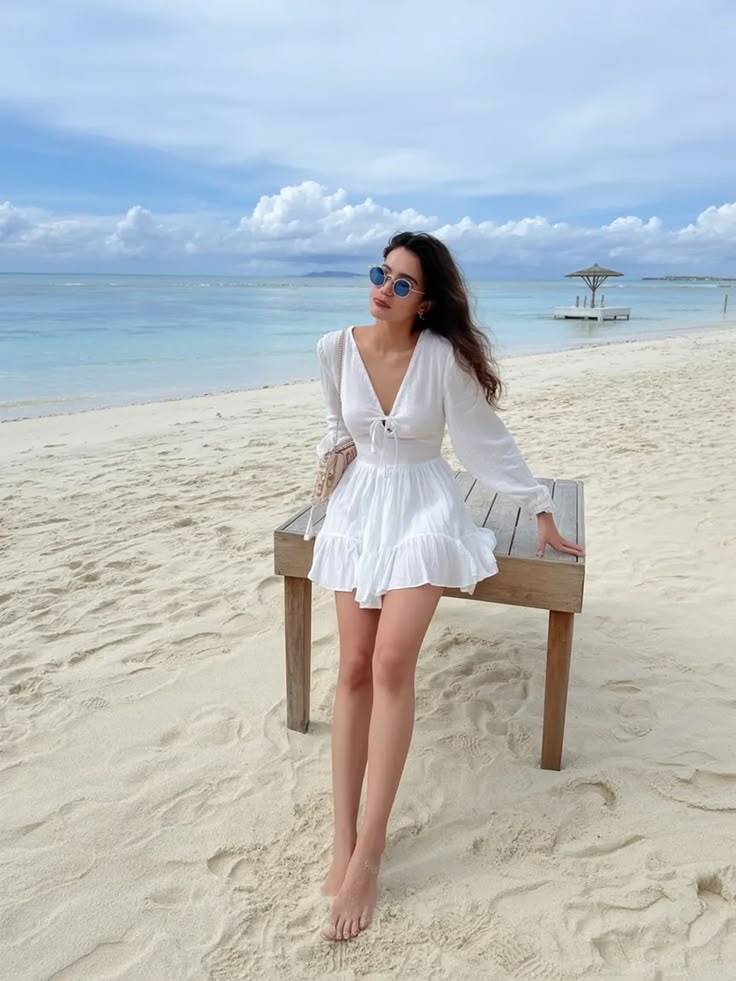 Woman in a white sundress with sunglasses leaning on a wooden bench on a sandy beach, calm turquoise sea in the background.