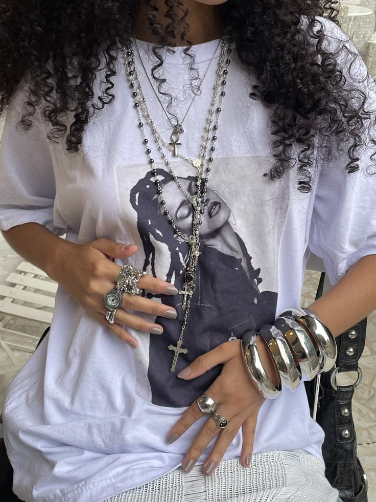 Person wearing layered silver necklaces with cross pendants over a white graphic tee, plus multiple rings and chunky bracelets.