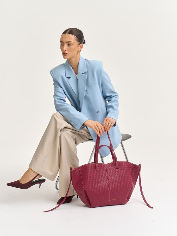 Woman seated on a chair wearing a light blue blazer and beige trousers, holding a large burgundy textured statement bags beside her.
