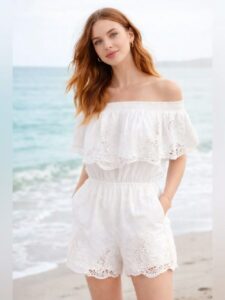Woman with long red hair wearing a white off-the-shoulder lace romper, standing on a beach with the sea in the background.