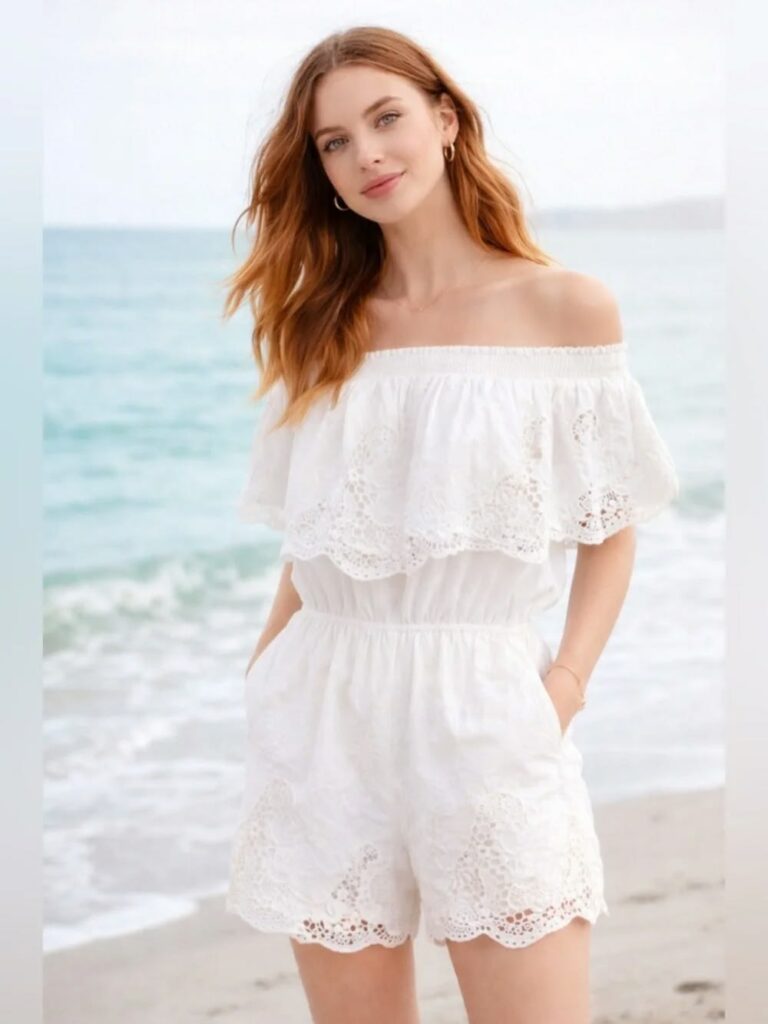Woman with long red hair wearing a white off-the-shoulder lace romper, standing on a beach with the sea in the background.