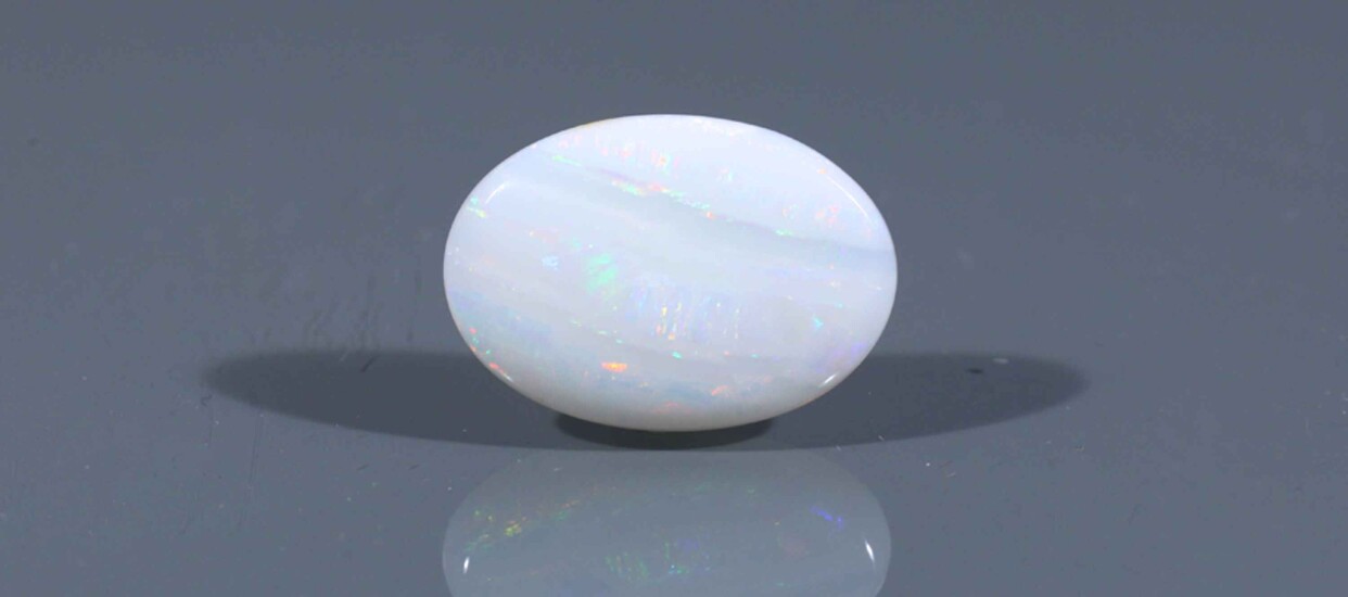 White October Birthstone with iridescent flashes resting on a gray surface with a soft shadow and reflection.