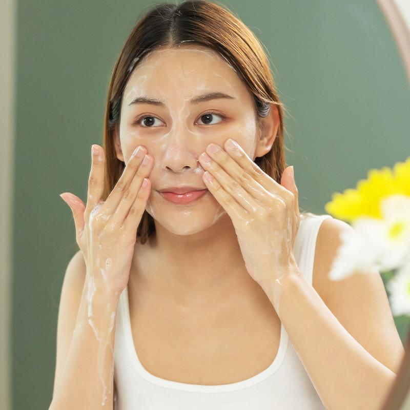 skincare routine - Close-up of a young woman washing her face with foamy cleanser, hands on her cheeks and looking into a mirror.