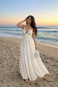 Woman in a white halter maxi dress walking barefoot on the sandy beach at sunset, holding a small white purse in front of her.
