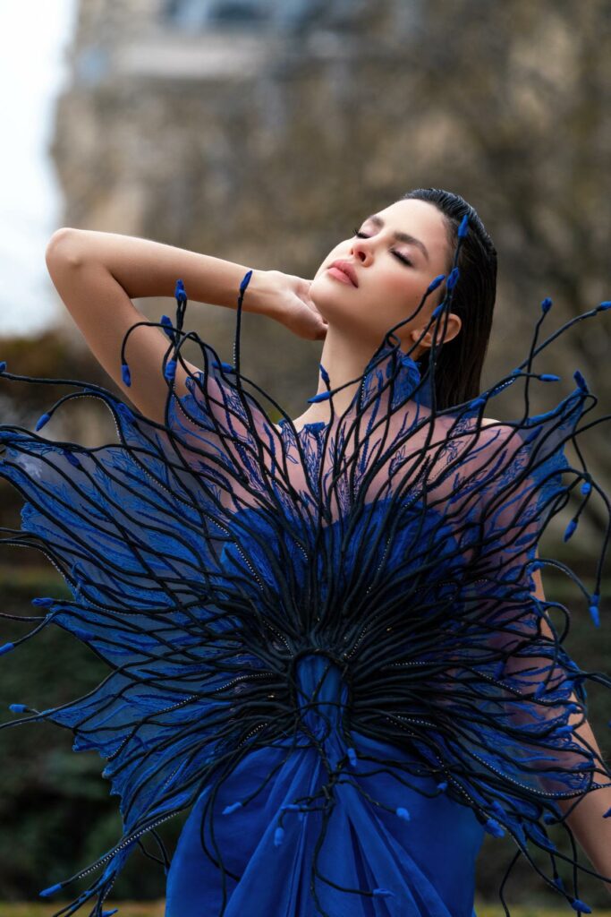 Portrait of a model in a vivid blue sculptural gown with dark blue tendrils; eyes closed, posing outdoors. Bio-Fabricated Character Dressing