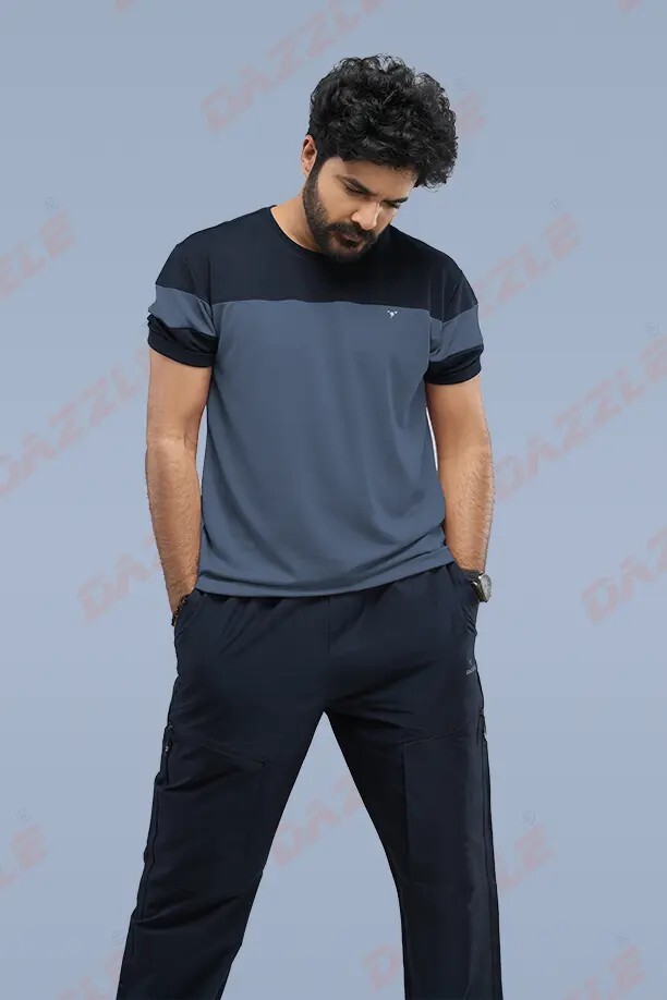 Full-body shot of a bearded man in a blue color-block T-shirt and black cargo pants, hands in pockets, looking down.