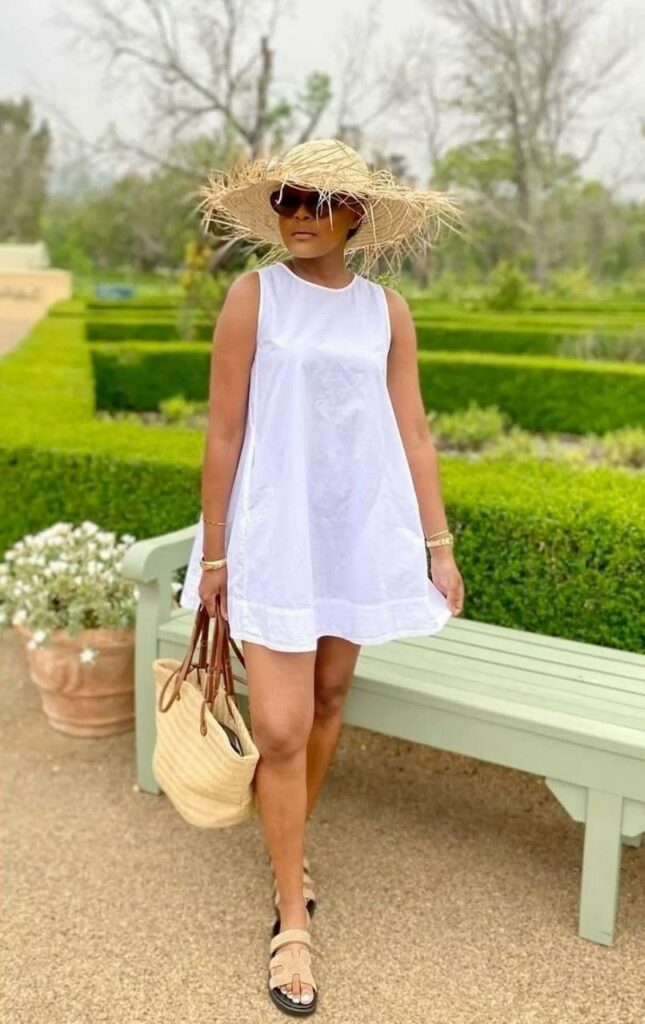 Woman in a white summer dress, straw hat, sunglasses, tan sandals, and a straw tote standing in a groomed garden.