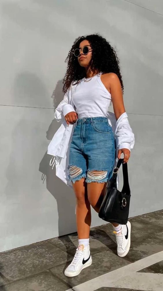 Woman with curly hair wearing a white tank top, ripped denim shorts, white sneakers, sunglasses, and a black handbag against a gray wall.