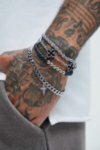 Tattooed forearm wearing layered silver bracelets, including a twisted chain, beaded black band, and cross pendant.