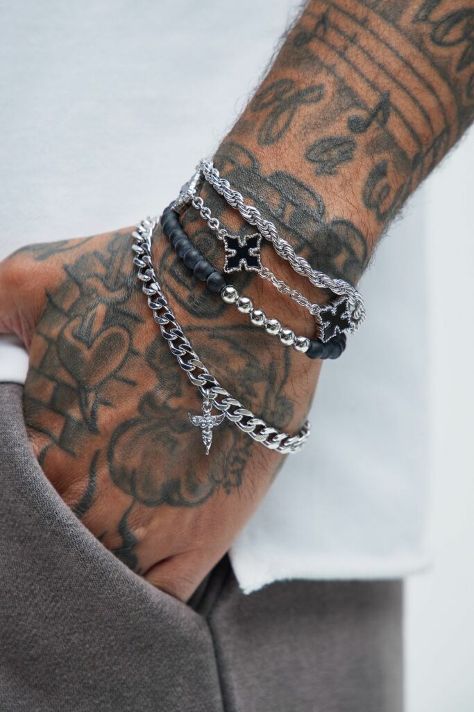 Tattooed forearm wearing layered silver bracelets, including a twisted chain, beaded black band, and cross pendant.