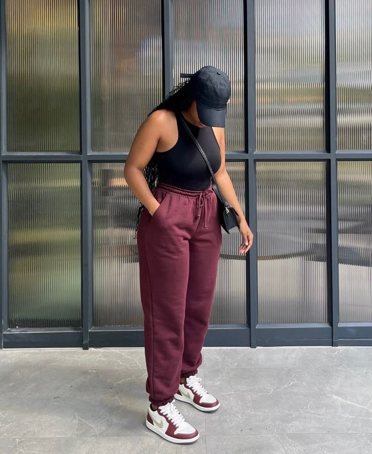 Person standing outdoors in front of a striped glass wall, wearing a black sleeveless top, burgundy sweatpants, white-and-burgundy sneakers, and a black cap with a crossbody bag.