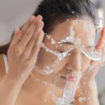 Woman washing her face with water splashing across her cheeks and eyes closed.