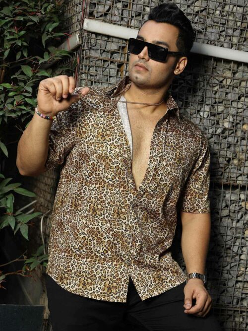 Man in a leopard print shirt with sunglasses, a gold chain, and bracelet, leaning against a wire fence with stones behind him.