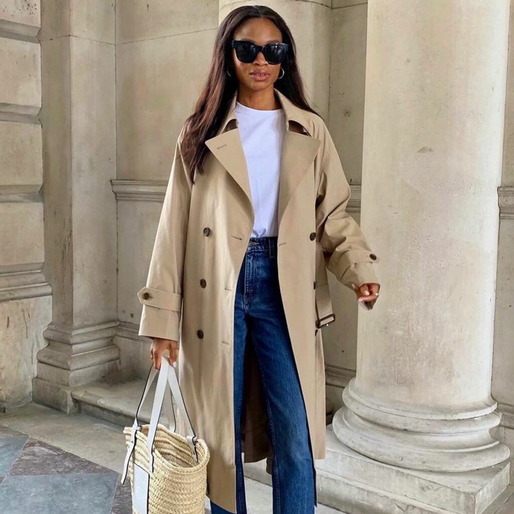 Woman in a beige trench coat, white T-shirt, and blue jeans, wearing black sunglasses and hoop earrings, carrying a straw tote near stone columns.