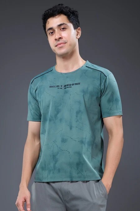 Man wearing a teal, marbled T-shirt with hands in pockets against a gray studio backdrop.
