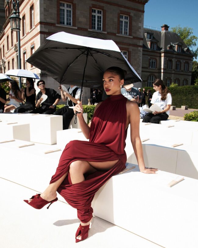 The design featured an elegant backless mock neck with ruching accents, flowing into a maxi skirt with matching ruching, a side slit, and finished with burgundy mules.
